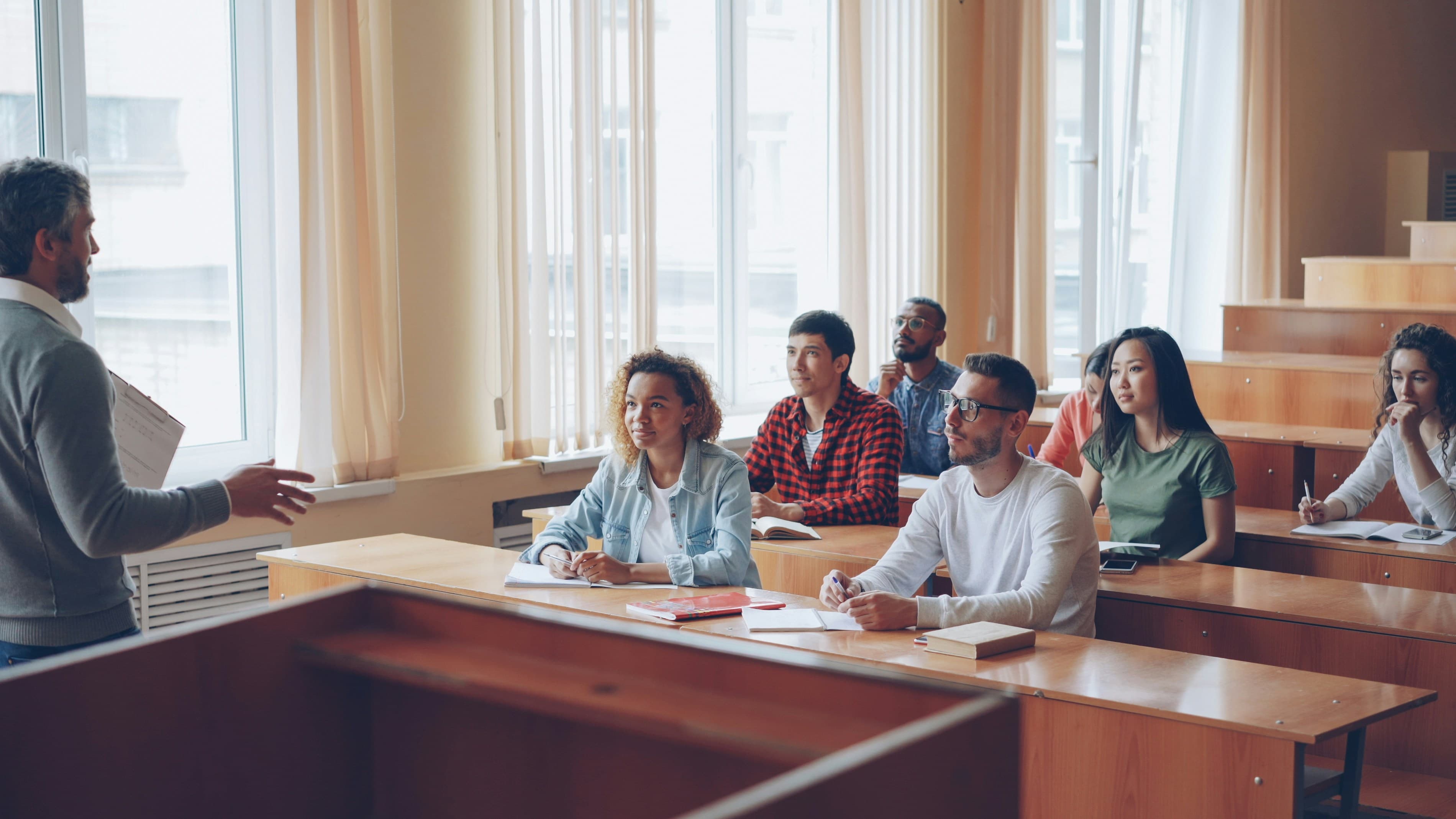 Students taking notes at tiered desks while an instructor leads a class in a sunlit lecture hall.